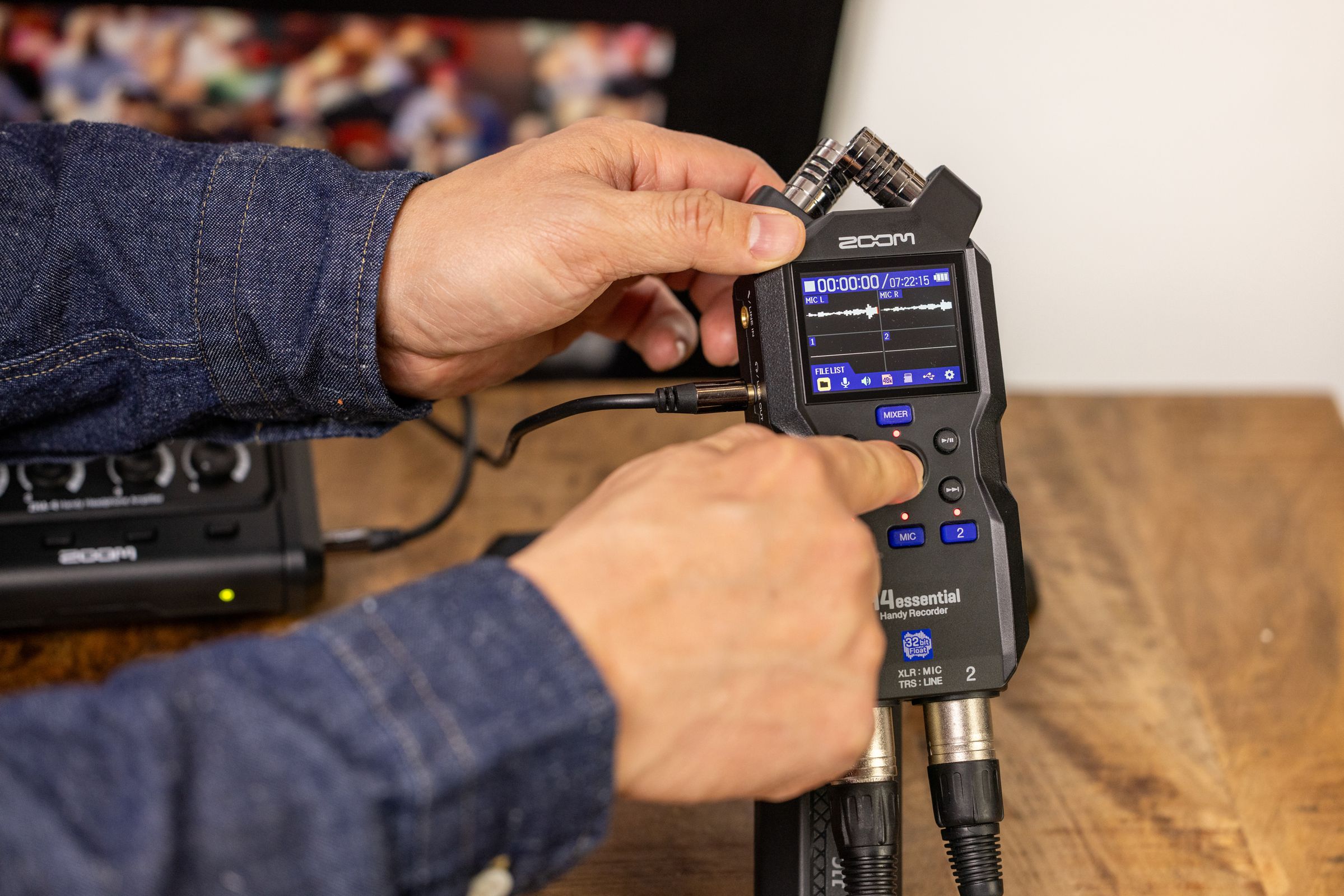 Zoom H4 Essential digital recorder with dual microphones and controls, shown in a lifestyle setting with headphones and audio equipment.