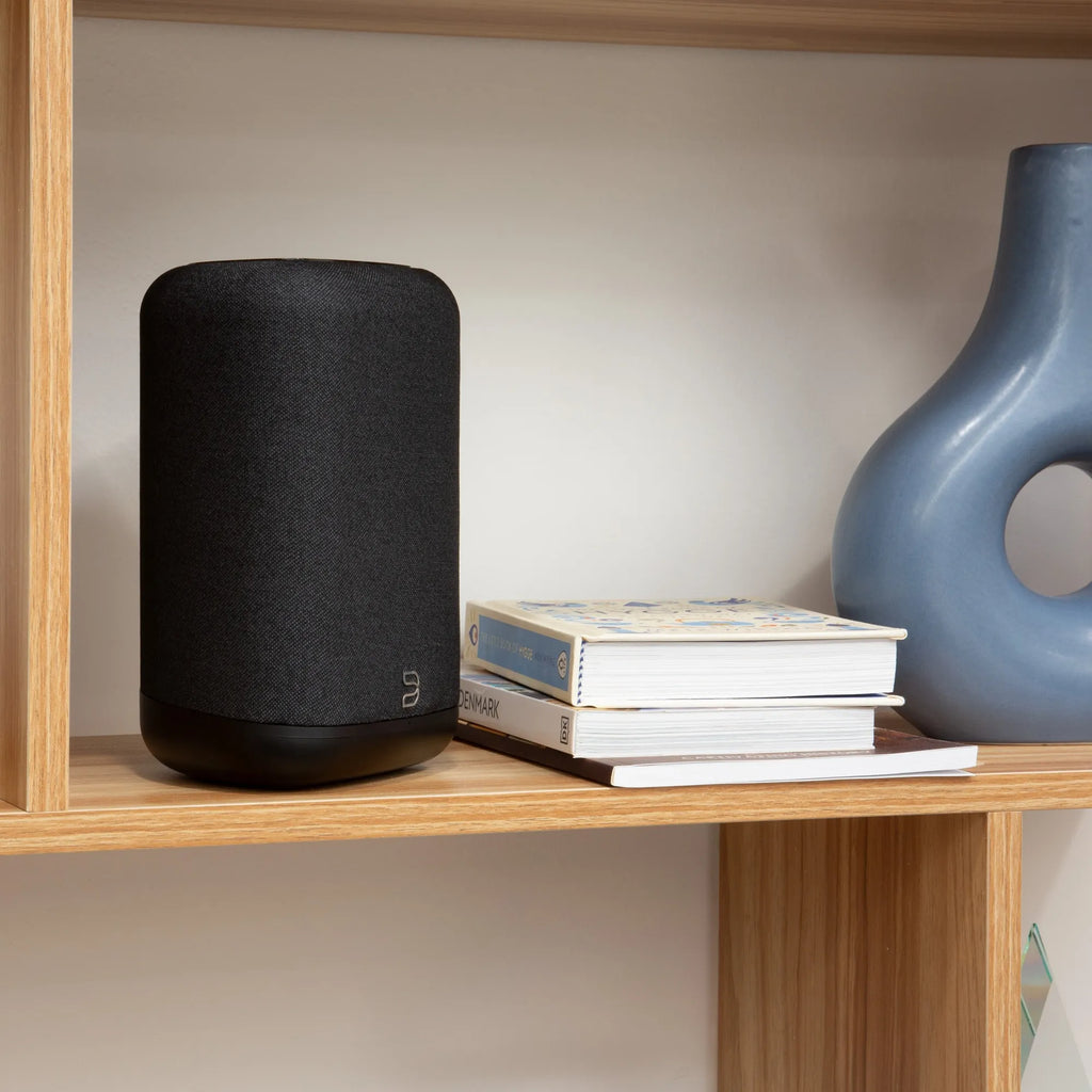 Black Bluesound Pulse Flex wireless speaker placed on a bookshelf with books, showcasing compact design and modern audio technology.