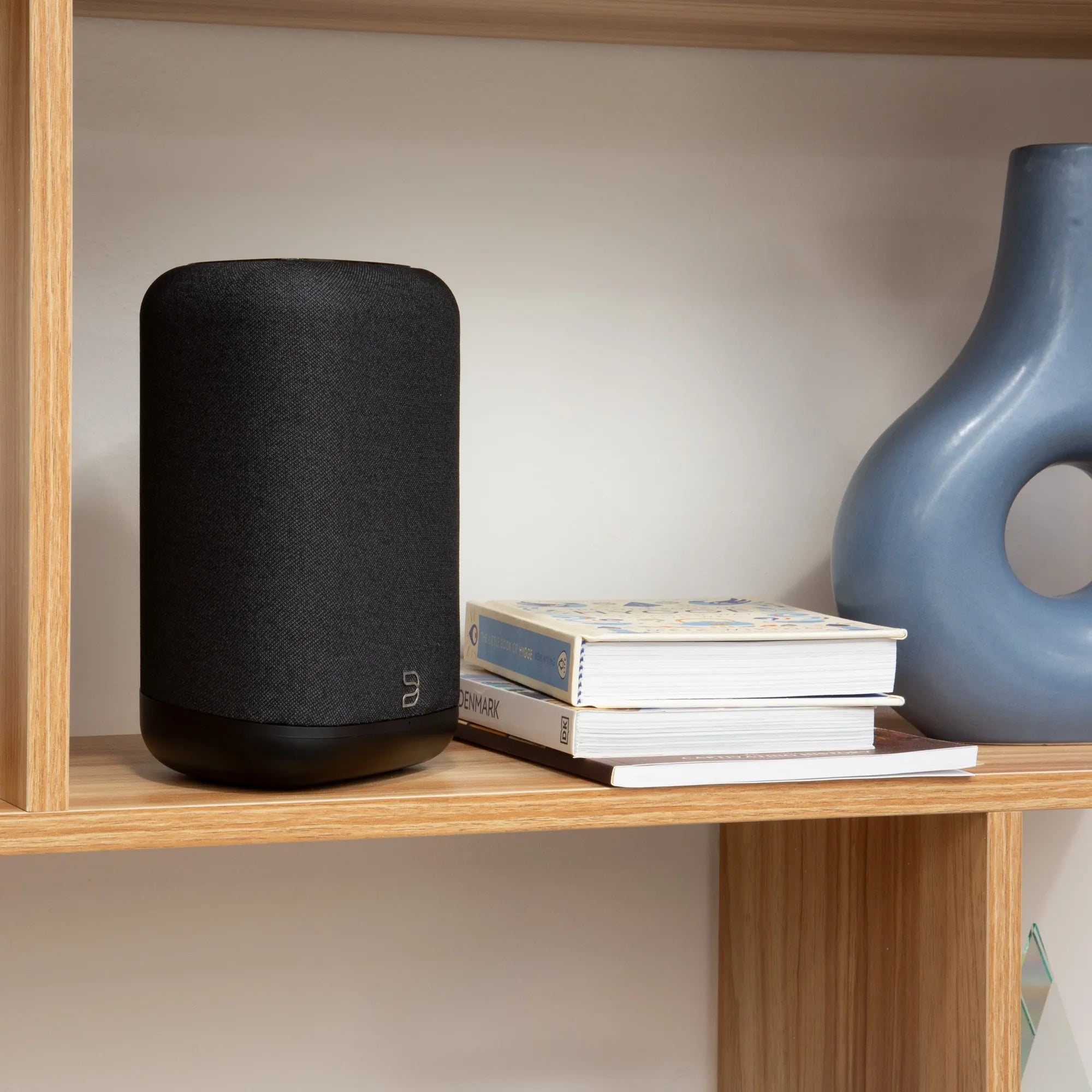 Black Bluesound Pulse Flex wireless speaker placed on a bookshelf with books, showcasing compact design and modern audio technology.
