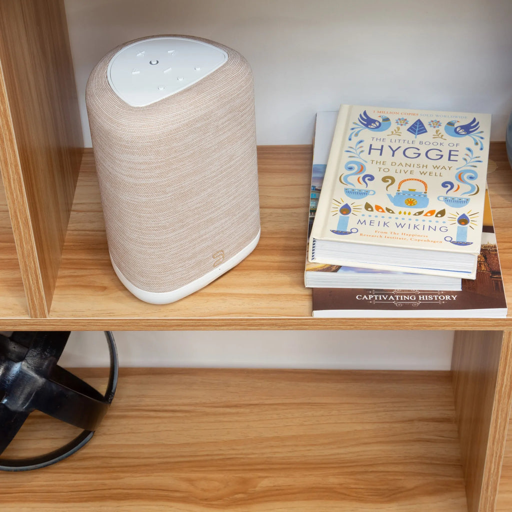 Bluesound Pulse Flex wireless speaker in tan, placed on a shelf above books, showcasing its compact design and modern look.