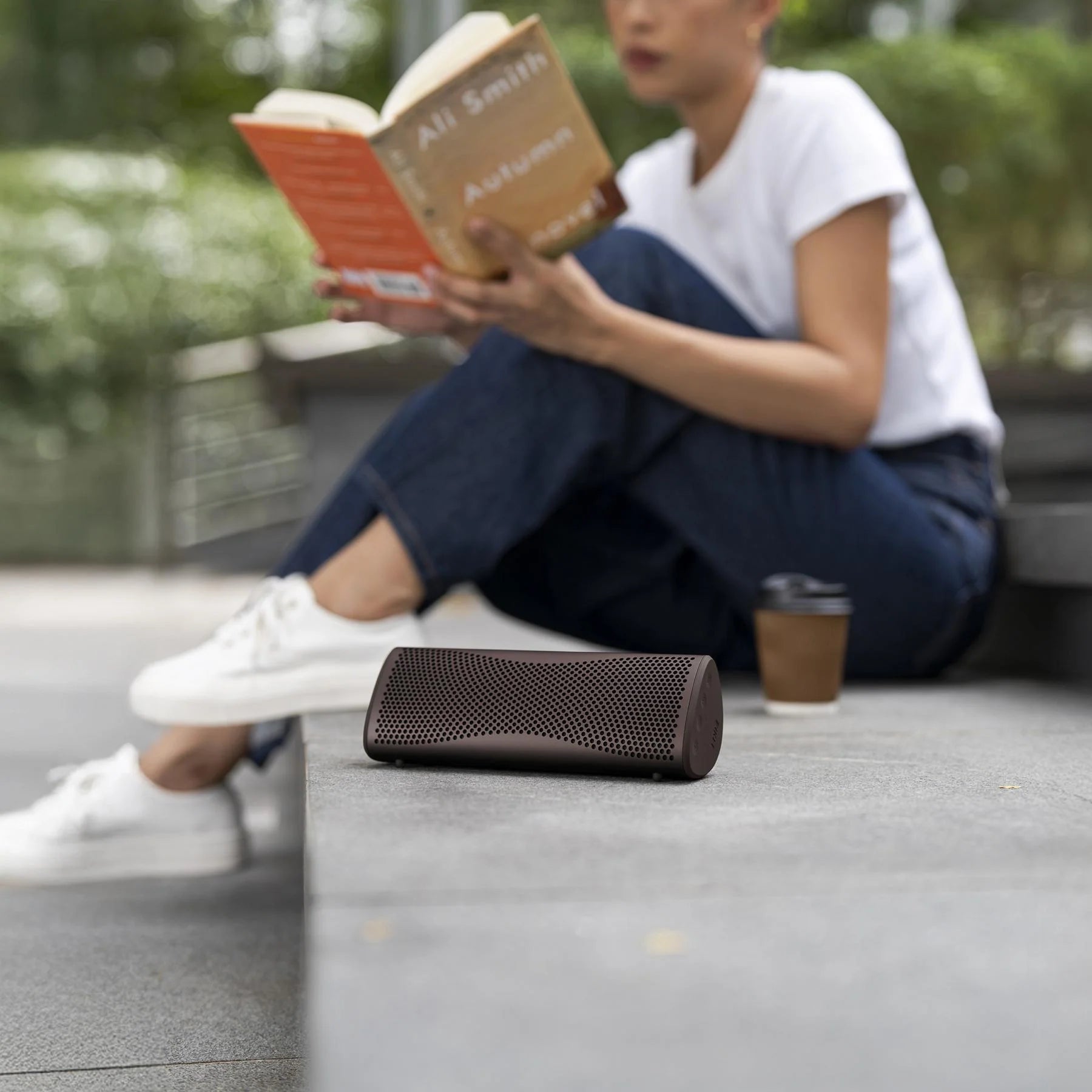 KEF Muo portable speaker in Cocoa Brown outdoors, showing compact design and fabric grille with control buttons.