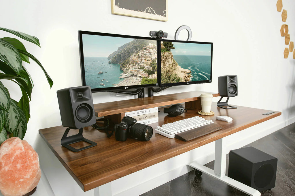 Kanto Audio Ora desktop speakers placed on a desk with a modern workspace setup, showing sleek black design and control knobs.